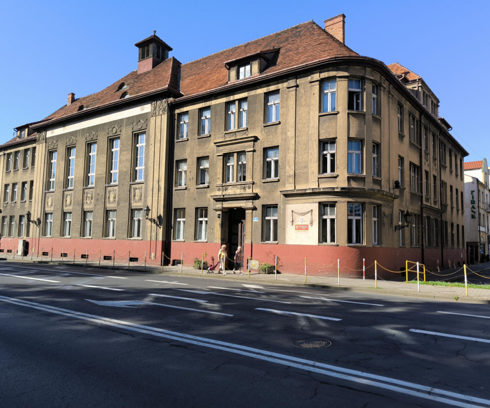 Budynek Szkoły Podstawowej nr 1 im. Marii Konopnickiej w Lesznie, obiekt z brązowym dachem i licznymi oknami, widoczny w słoneczny dzień. Przed szkołą znajduje się ulica z pasami dla pieszych i dwoje przechodniów. Więcej o przyszłości placówki przeczytasz na naszym portalu.
