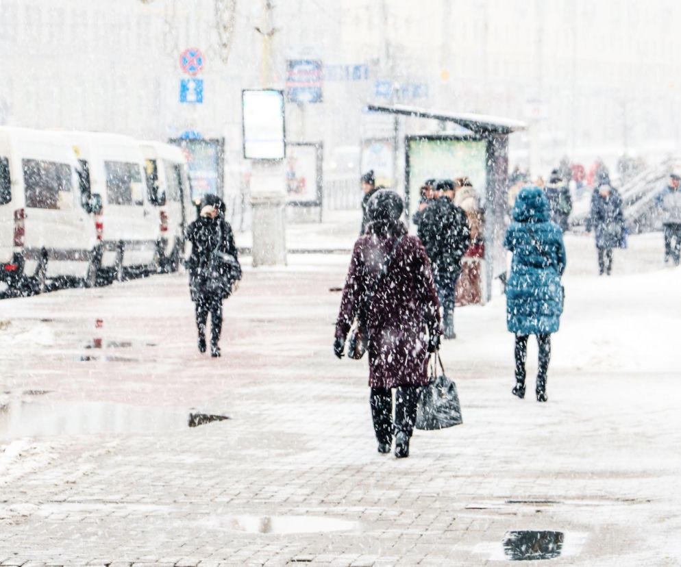 Ludzie idący chodnikiem podczas obfitych opadów śniegu. Ubrani w ciepłe, zimowe kurtki, starają się pokonać śliską nawierzchnię. W tle widać ulicę z zaparkowanymi samochodami dostawczymi oraz przystanek autobusowy. O surowej zimie przeczytasz na Poradnik Zdrowie.