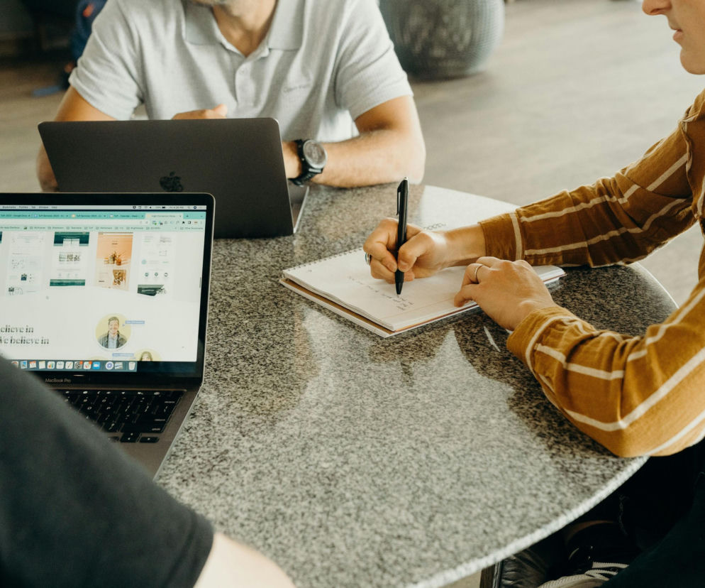 Trzy osoby przy stoliku, jedna pracuje na laptopie z otwartą stroną internetową, inna robi notatki, a trzecia siedzi z tyłu. Scena obrazuje analizę wiarygodności kontrahenta, o której przeczytasz na Super Biznes.