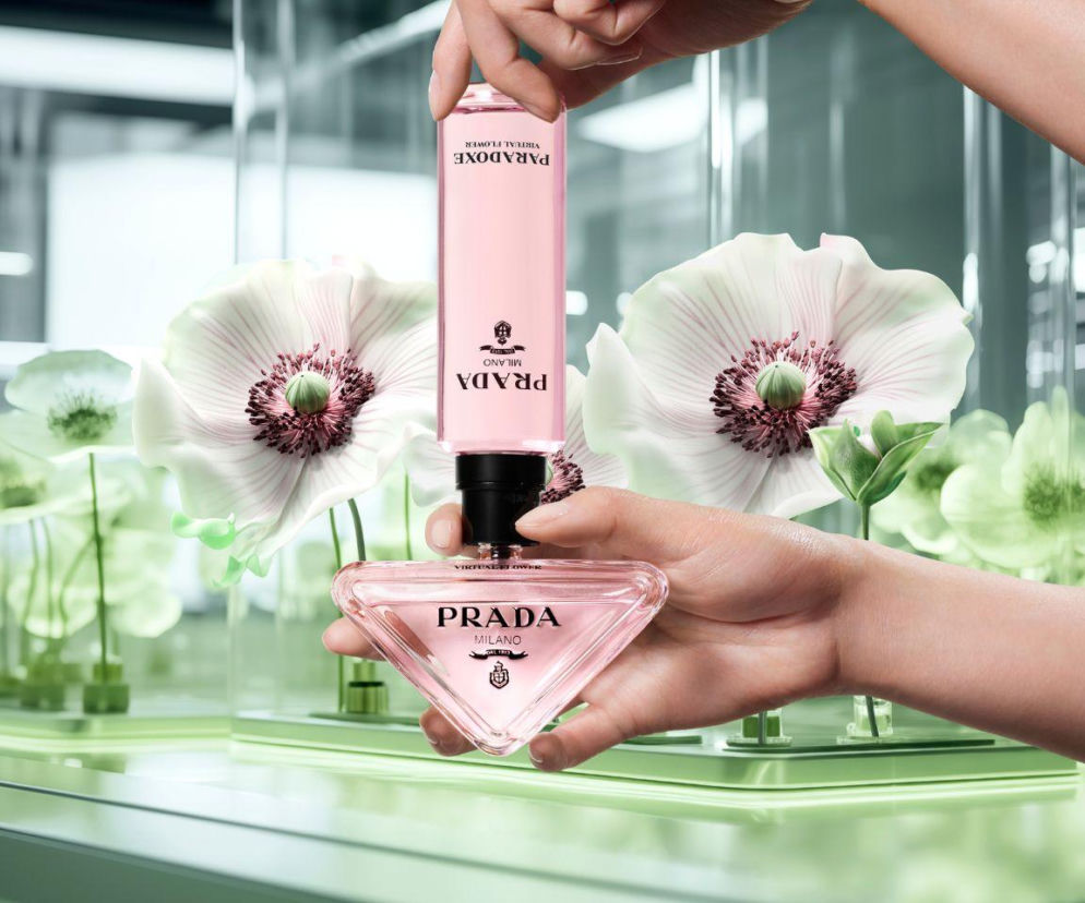Dwie dłonie uzupełniają flakon perfum Prada Paradoxe różowym płynem z refilla, symbolizując zrównoważone wybory konsumenckie. Tło stanowią delikatne białe kwiaty, podkreślające estetykę beauty. Więcej o trendzie refill przeczytasz na naszym portalu.