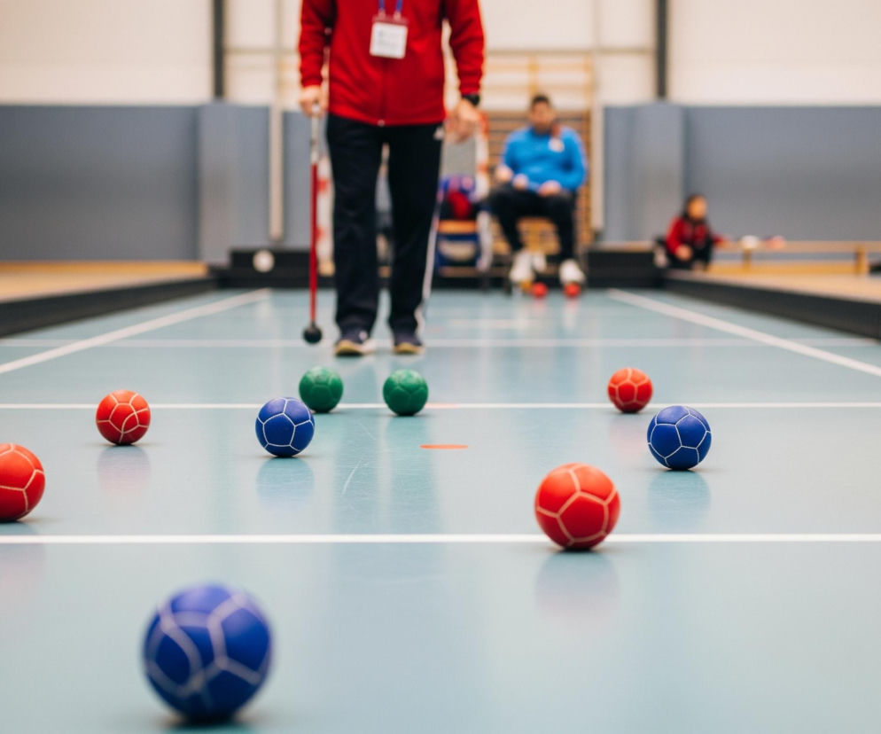 Na boisku sportowym o jasnoniebieskiej nawierzchni, podzielonym białymi liniami, leży wiele kolorowych piłek, głównie czerwonych, niebieskich i zielonych, rozmieszczonych w losowy sposób. W centrum, lekko z tyłu, stoi osoba w czerwonej bluzie i ciemnych spodniach, trzymająca długi czerwony kij z czarną końcówką, patrząca w kierunku piłek. W tle widać rozmyte sylwetki innych osób, niektóre siedzące na ławkach lub krzesłach, oraz ścianę hali sportowej.