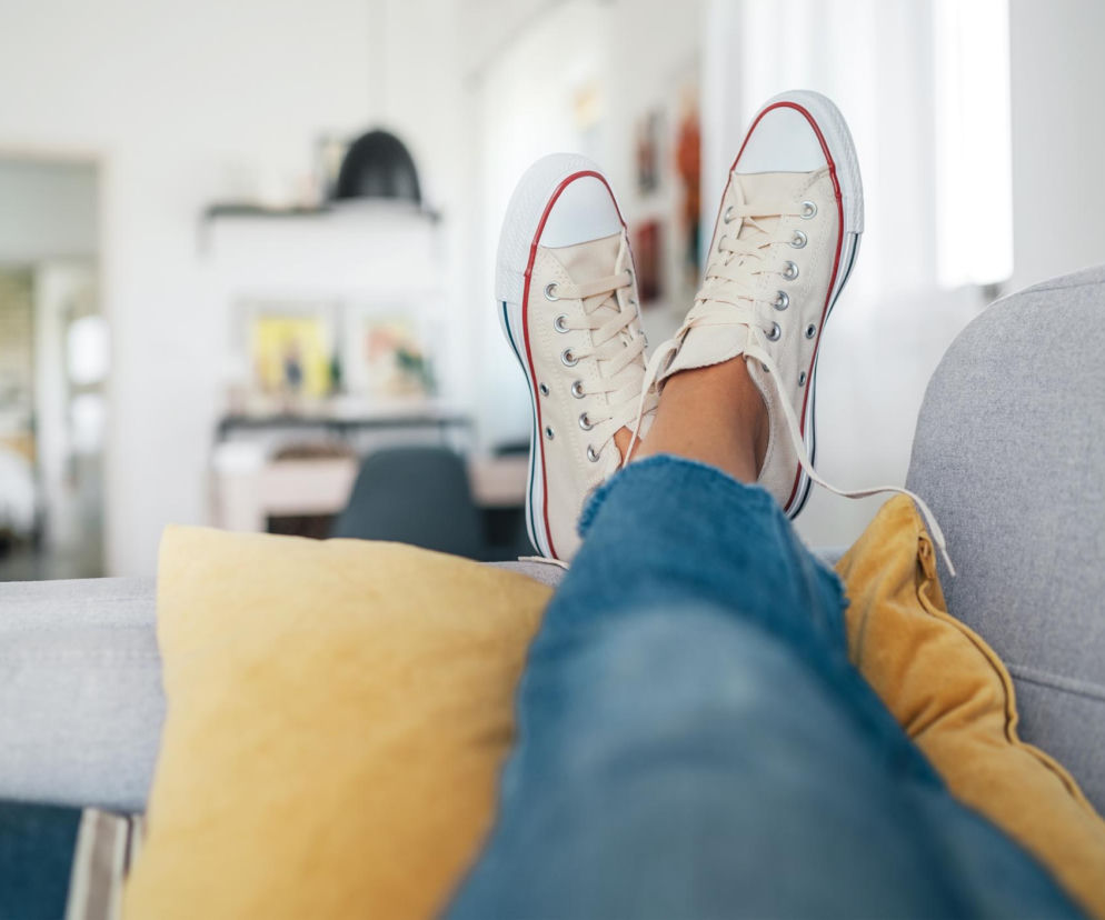 Osoba w dżinsach leży na szarej sofie z nogami wspartymi na żółtej poduszce, ubrana w jasne tenisówki z czerwonymi akcentami. Stopa w obuwiu jest blisko obiektywu, a za nią widać rozmyte wnętrze pokoju, ilustrując temat wnoszenia bakterii do domu na butach, o czym przeczytasz na Poradnik Zdrowie.