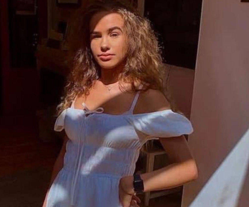 Młoda influencerka Brooke George w białej sukience z odkrytymi ramionami, z długimi, kręconymi włosami. Jest to zdjęcie portretowe, na którym influencerka pozuje w pomieszczeniu, oświetlona częściowo przez promienie słońca. Więcej o tej szokującej sprawie przeczytasz na naszym portalu.