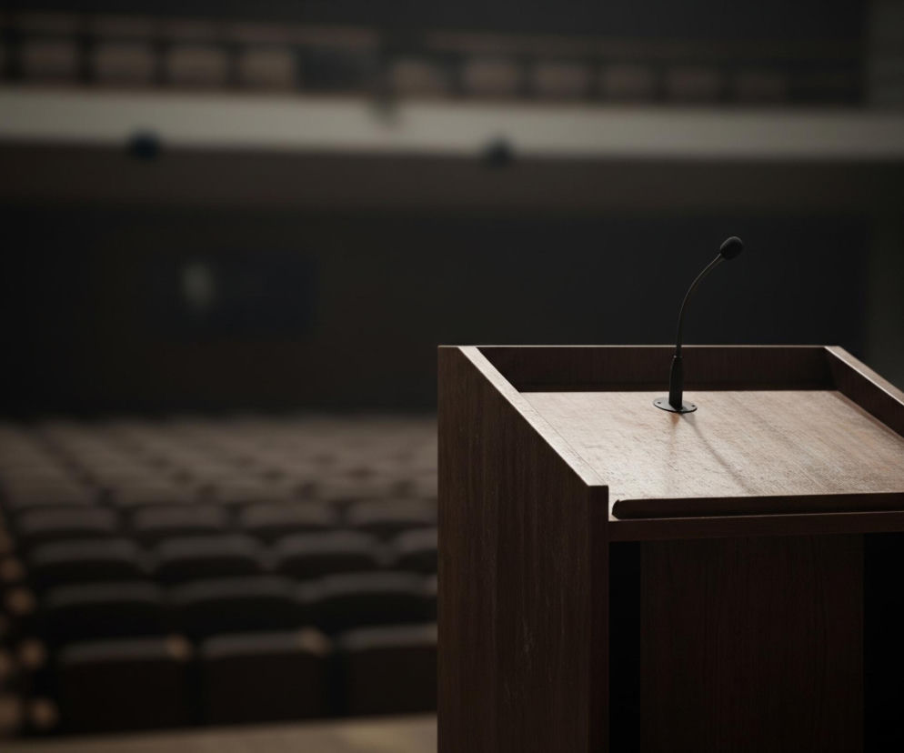 Na pierwszym planie, z prawej strony, znajduje się drewniana mównica o ciemnobrązowej barwie, z subtelnym usłojeniem. Na jej płaskiej powierzchni umieszczony jest czarny mikrofon na giętkiej szyjce, skierowany w lewo. Tło przedstawia pustą salę wykładową lub audytoryjną, z rzędami ciemnych foteli ustawionych symetrycznie. Cała scena jest słabo oświetlona, co nadaje jej stonowany i poważny charakter, a większość tła jest rozmyta.