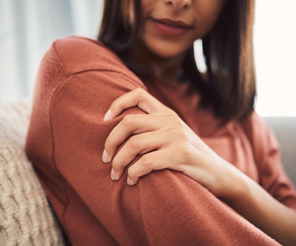 Młoda kobieta w rudym swetrze chwyta się za bolące ramię, czując nagłe, ostre ukłucie, co jest częstym, lecz niepokojącym objawem, o którym warto przeczytać na portalu Poradnik Zdrowie, by dowiedzieć się, kiedy należy skonsultować się z lekarzem.