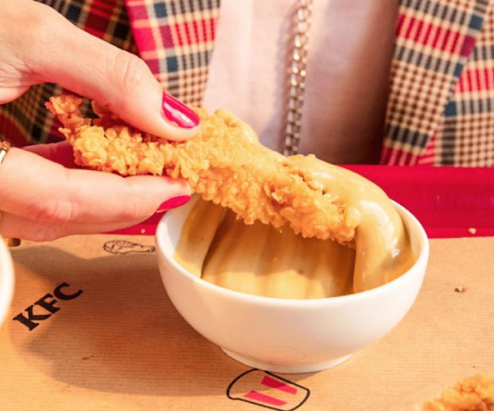 Dłoń kobiety maczającej kurczaka KFC w sosie. O powrocie wtorkowego kubełka do KFC przeczytasz na SE.