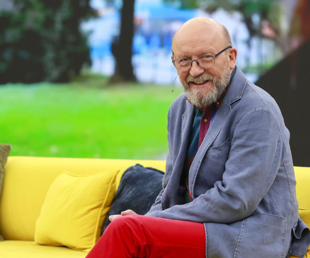 Uśmiechnięty aktor Artur Barciś w okularach i siwej brodzie siedzi na żółtej kanapie. O anegdotach aktora przeczytasz na SE.