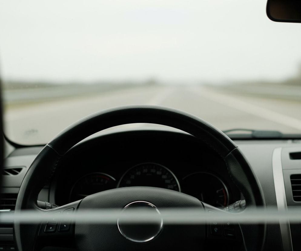 Czarne wnętrze samochodu jest widoczne z perspektywy pasażera. Na pierwszym planie po lewej stronie znajduje się czarny zagłówek fotela, częściowo zasłaniający widok na deskę rozdzielczą. Centralnym elementem jest czarna kierownica ze srebrnym okręgiem w środku, a za nią widoczne są zegary, w tym prędkościomierz. Po prawej stronie widać fragment deski rozdzielczej z kratkami nawiewu i czerwonym przyciskiem ostrzegawczym. Przednia szyba ukazuje szarą, lekko zamgloną drogę z biało-szarymi pasami, otoczoną niewyraźnymi elementami krajobrazu, a u góry lusterko wsteczne.