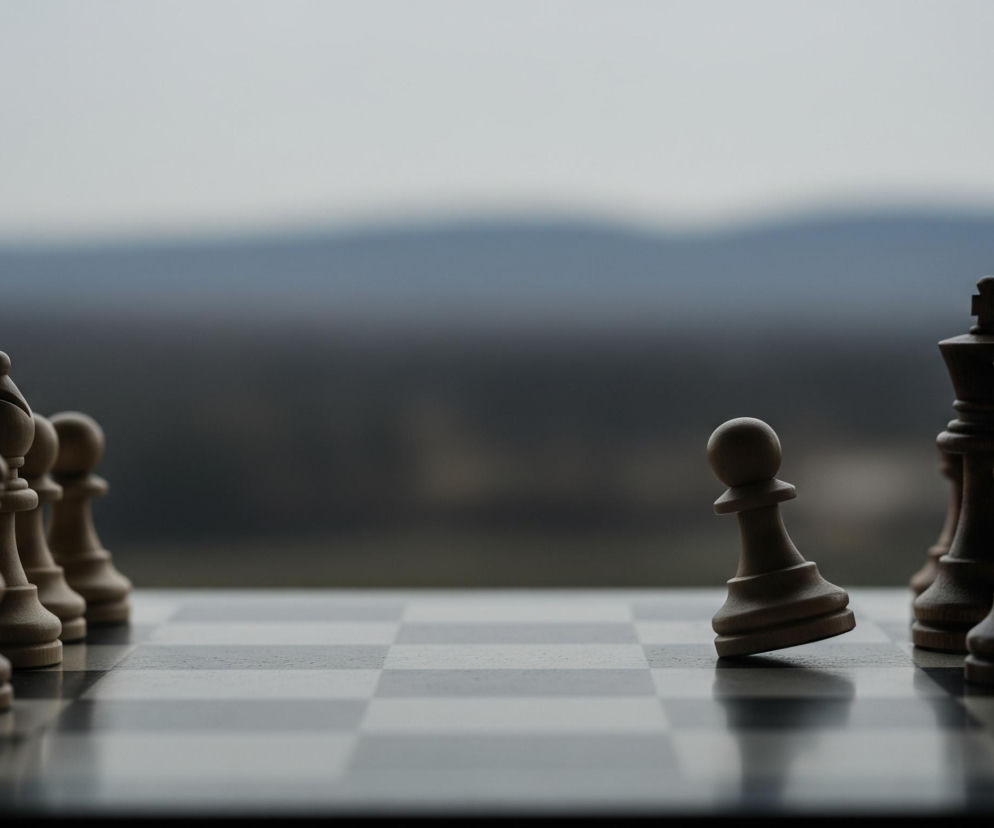Na pierwszym planie widoczna jest szachownica z figurami szachowymi. Jasnobrązowy pionek przechyla się w prawo, znajdując się blisko centrum zdjęcia. Po lewej stronie, w pierwszym rzędzie, stoi rząd jasnych figur szachowych, a po prawej stronie ciemniejsze figury szachowe. Tło stanowi rozmyty krajobraz z ciemnymi wzgórzami u dołu i jaśniejszym, szaroniebieskim niebem u góry.