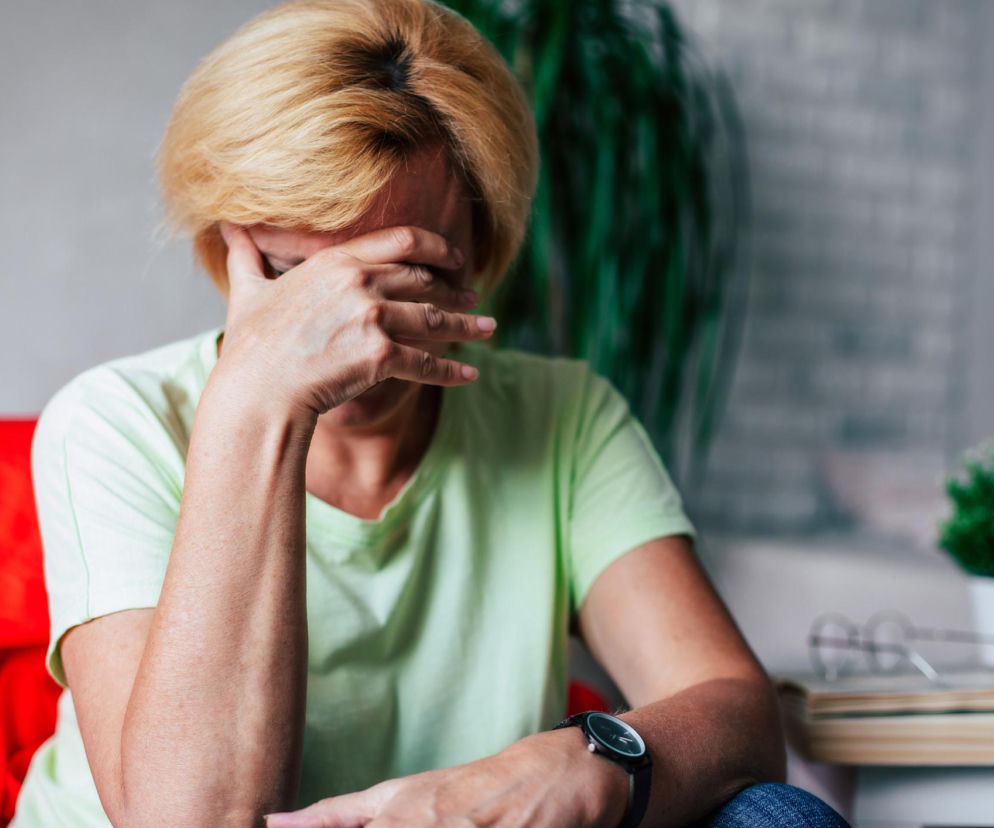 Zmęczona kobieta z jasnymi włosami, w zielonej koszulce, siedząca na czerwonej kanapie, zakrywa twarz dłonią, sygnalizując chroniczne zmęczenie. W tle rozmyte rośliny i książki. Na portalu Poradnik Zdrowie znajdziesz więcej informacji o wpływie cichego hałasu na zdrowie.