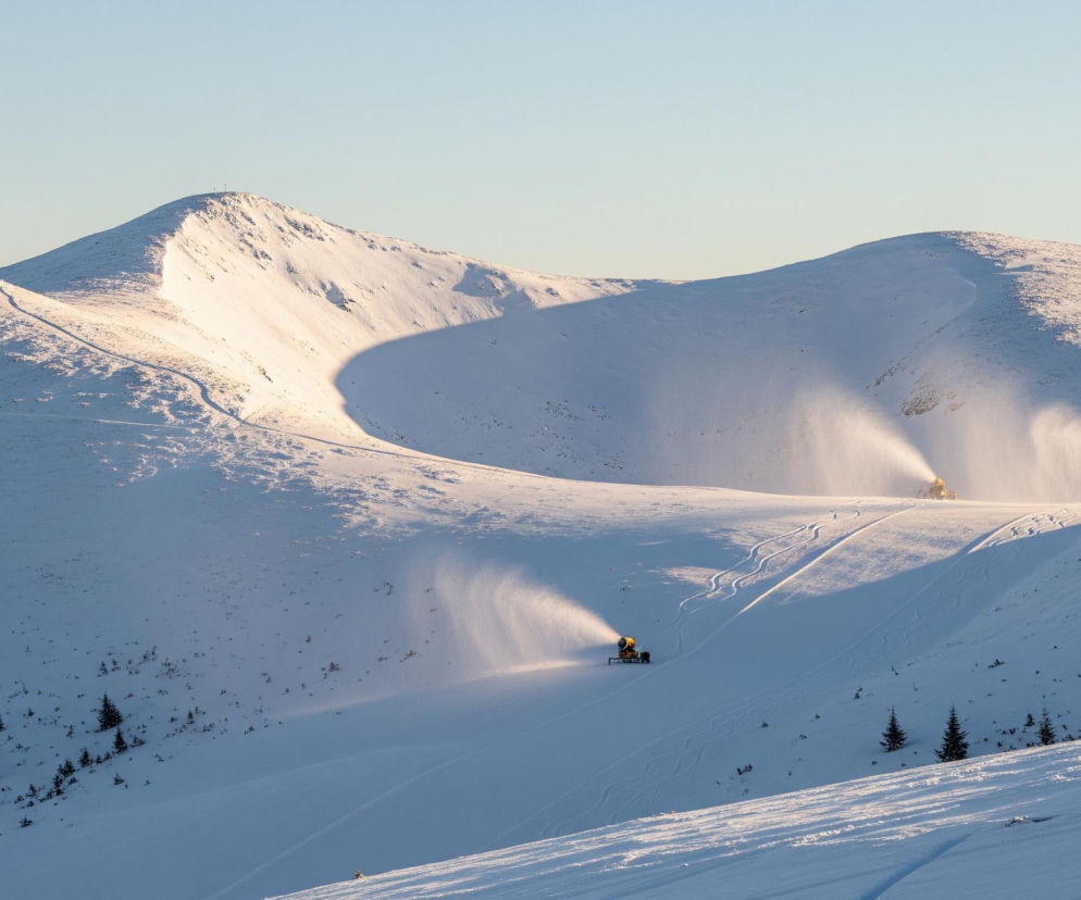 Trzy żółte armatki śnieżne są rozmieszczone na zaśnieżonym stoku, aktywnie produkując sztuczny śnieg, który unosi się w powietrzu, tworząc smugi białego puchu. Górna część obrazu ukazuje rozległe, pokryte śniegiem góry, z których jedna, położona w lewej części, ma wyraźny szczyt z kilkoma drobnymi obiektami na wierzchołku, prawdopodobnie masztami. Śnieg mieni się w ciepłym świetle wschodzącego lub zachodzącego słońca, rzucając długie cienie na pofalowany teren. Dolna część kadru przedstawia fragment zbocza pokrytego rzadkim lasem iglastym, a na stoku widać ślady nart lub innych pojazdów śnieżnych.