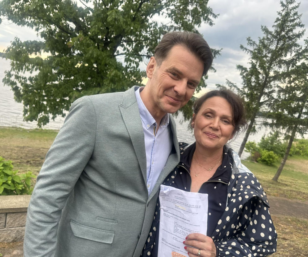 Aktor Marcin Kwaśny (jako Jerzy Wielicki) i aktorka Małgorzata Pieńkowska (jako Marysia) uśmiechają się do zdjęcia na tle jeziora i zielonych drzew. Małgorzata trzyma scenariusz serialu. Zdjęcie ilustruje newsy z portalu Super Seriale.