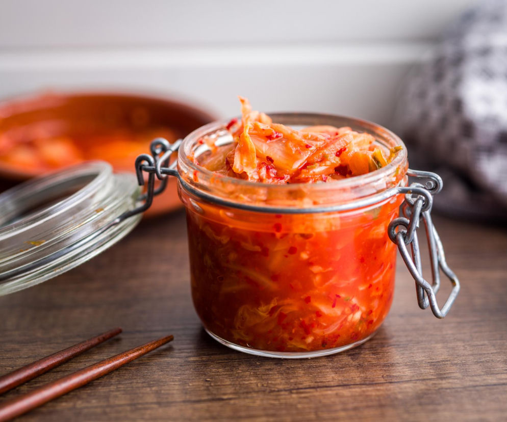 Słoik z otwartą pokrywką pełen czerwonego, pikantnego kimchi z kawałkami kapusty pekińskiej, stoi na drewnianym stole obok pałeczek. Z tyłu widać ceramiczną miskę z kimchi. To smaczny produkt, o którego wpływie na zdrowie przeczytasz na portalu Poradnik Zdrowie.