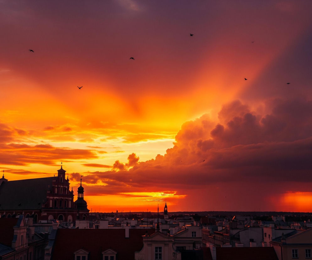 Pano rama miejskiego krajobrazu o zachodzie słońca ukazuje miasto skąpane w intensywnych barwach pomarańczy i czerwieni. Po lewej stronie dominuje fasada budynku w odcieniach beżu i brązu z wyraźnymi oknami, stanowiąca ramę dla widoku. Na środku, na tle purpurowo-czerwonego nieba, wyróżnia się sylwetka dużego, ciemnego kościoła z wysokim dachem i wieżyczkami. Horyzont miasta jest usiany niskimi budynkami o jasnych elewacjach i czerwonych dachach, a na ciemnoczerwonym niebie widoczne są rozproszone ciemne chmury, z których po prawej stronie pada deszcz. Liczne ptaki w locie są widoczne na tle jaskrawego zachodzącego słońca, które rzuca długie, pomarańczowe smugi światła przez chmury.