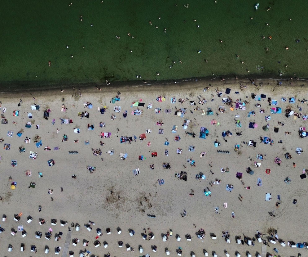 Plaża pełna ludzi wypoczywających nad zieloną wodą, widziana z lotu ptaka. Wiele kolorowych ręczników i parasoli świadczy o upalnym dniu. To ilustruje rekordowe temperatury w Europie, o których przeczytasz więcej na naszym portalu.