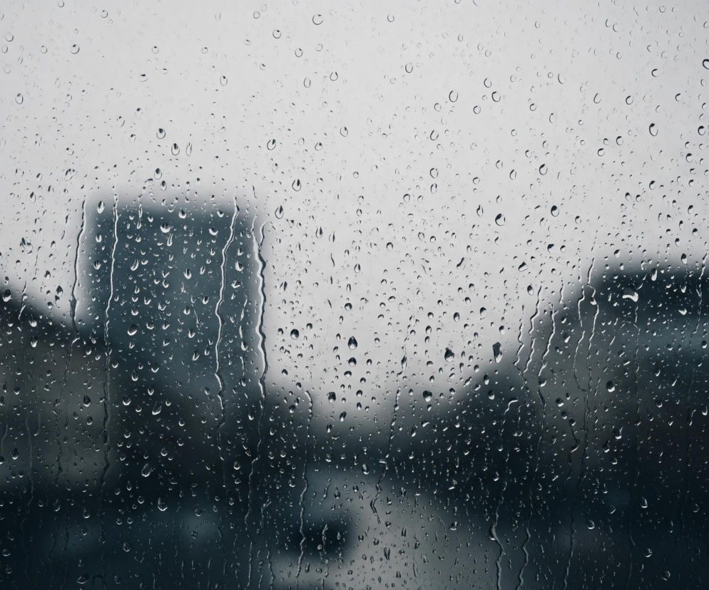 Powierzchnia szyby jest pokryta licznymi kroplami wody, o różnej wielkości i kształcie, niektóre z nich zlewają się, tworząc smugi spływające pionowo w dół. Widok zza szyby jest rozmyty i nieostry, dominują ciemne, szare i niebieskawe odcienie. Na dalszym planie, w centrum, widoczna jest niewyraźna, ciemna sylwetka budynku, a po jego bokach zarys innych budynków lub drzew. U dołu kadru, w środkowej części, przebiega rozmazana, ciemniejsza linia, przypominająca jezdnię z niewyraźnym kształtem samochodu.