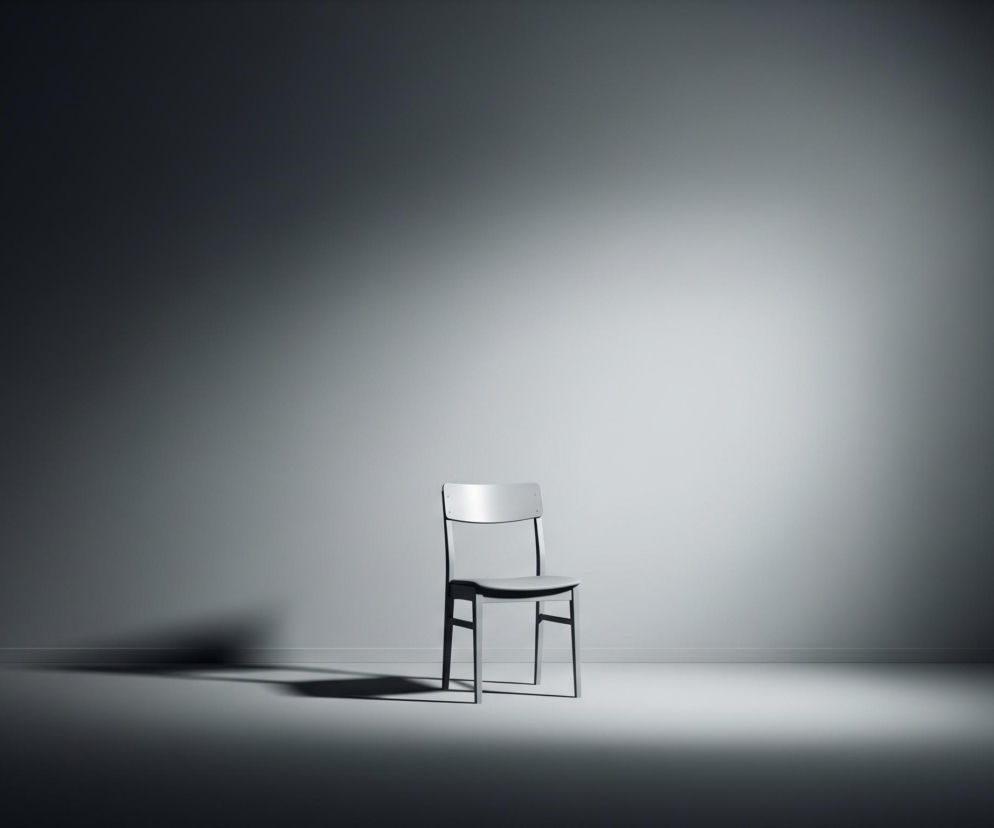 Centralnie umieszczone jest proste krzesło o jasnoszarym siedzisku i oparciu, z ciemnymi, cienkimi nogami. Stoi ono na gładkiej, szarej podłodze, która łączy się z równie gładką ścianą w tle, tworząc minimalistyczne wnętrze. Całość oświetlona jest silnym, centralnym światłem padającym od przodu, rzucającym długi, rozmazany cień krzesła na podłogę po lewej stronie. Światło tworzy gradację tonów od jasnego centrum do ciemnych, niemal czarnych krawędzi obrazu.
