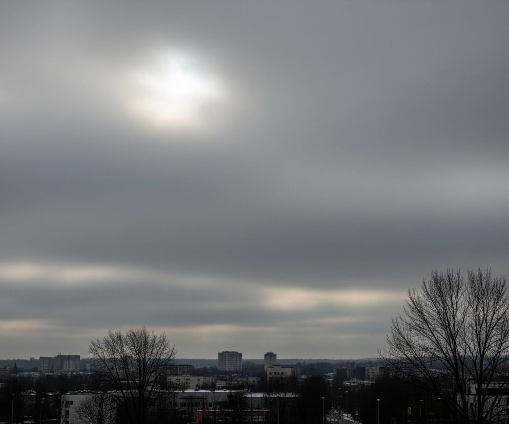 Niebo wypełnione jest gęstymi, szarymi chmurami, z jasną, rozmazaną plamą światła słonecznego przebijającego się przez centrum górnej części kadru. Poniżej, na horyzoncie, rozciąga się panorama miasta z licznymi budynkami mieszkalnymi o różnych kształtach i wysokościach, otoczonymi nagimi drzewami bez liści. Cała scena utrzymana jest w chłodnych, stonowanych odcieniach szarości i brązu, sugerując pochmurny, zimowy dzień.