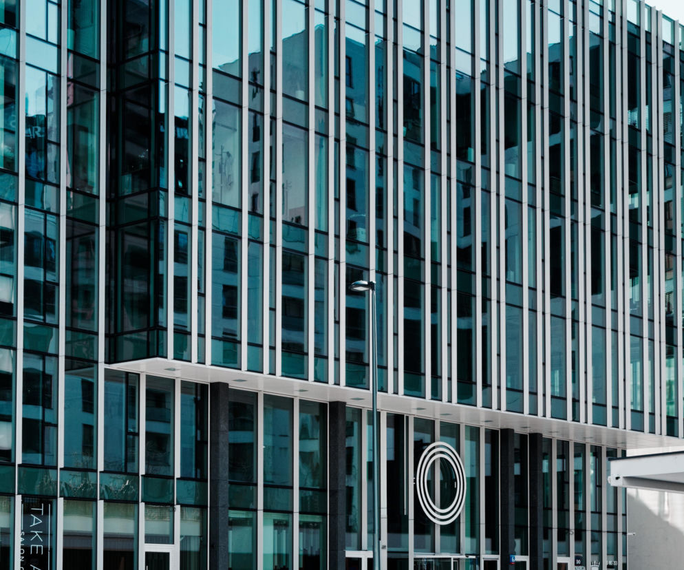 Nowoczesny biurowiec MetroPoint Office przy Hrubieszowskiej 6b w Warszawie, zaprojektowany przez FS&P Arcus Sp. z o.o., z elegancką szklaną elewacją i pionowymi żyletkami. Na dole dwie osoby przechodzące przez ulicę. Więcej o projekcie na Architektura Murator Plus.