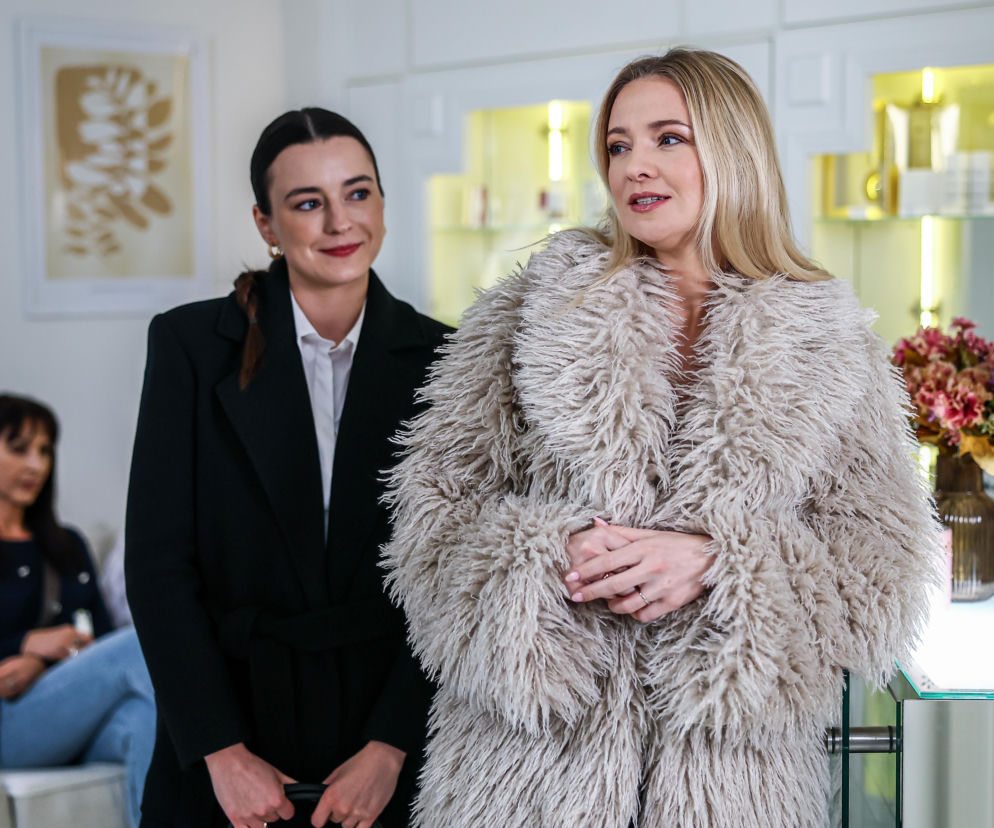 Blondwłosa Katarzyna Hajduk, w którą wciela się Joanna Liszowska, w modnym futerku rozmawia z prawniczką. W tle recepcja salonu. Na Super Seriale znajdziesz więcej informacji o serialu Szpital św. Anny i losach Kaśki.