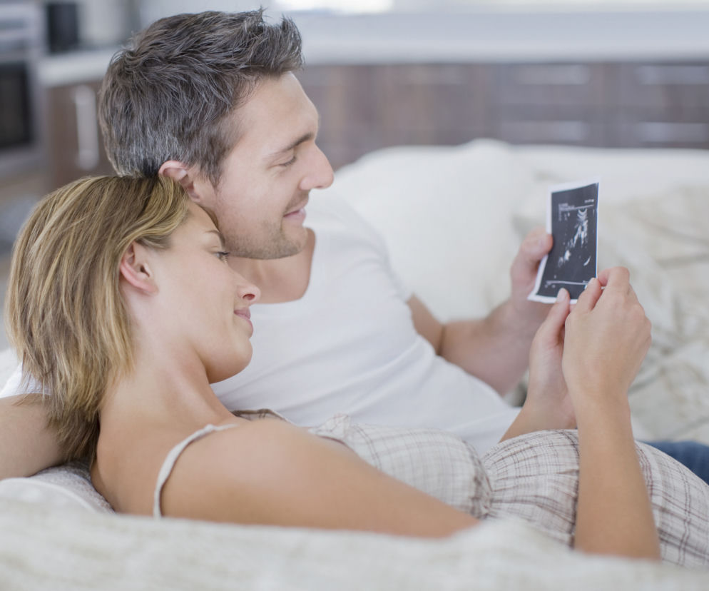 Uśmiechnięta para leży razem na kanapie, przytulając się. Kobieta trzyma w ręku zdjęcie USG ciąży i pokazuje je mężczyźnie. Oboje z radością patrzą na obraz. Więcej o objawach ciąży u mężczyzn przeczytasz na portalu Poradnik Zdrowie.