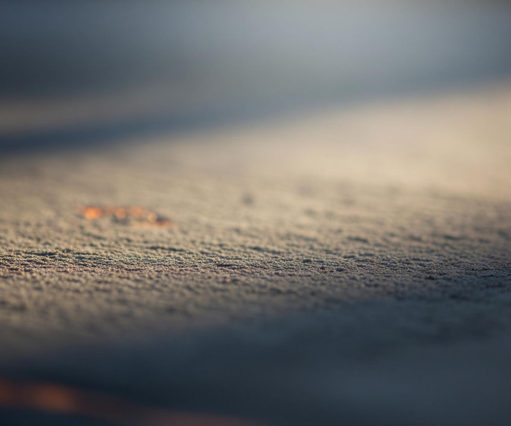 Powierzchnia pokryta jest drobnymi, szarymi granulkami, przypominającymi szron lub piasek, które częściowo oświetla jasne, ciepłe światło z prawej strony. Centralny obszar jest ostry, ukazując detale tekstury, natomiast górna i dolna część obrazu są rozmyte. W górnej części widać ukośną, ciemniejszą strefę, tworzącą cień, oraz jaśniejszy, mleczno-beżowy obszar. W dolnej części obrazu dominują ciemne barwy, z delikatnym, rozmytym pomarańczowym pasmem po lewej stronie.