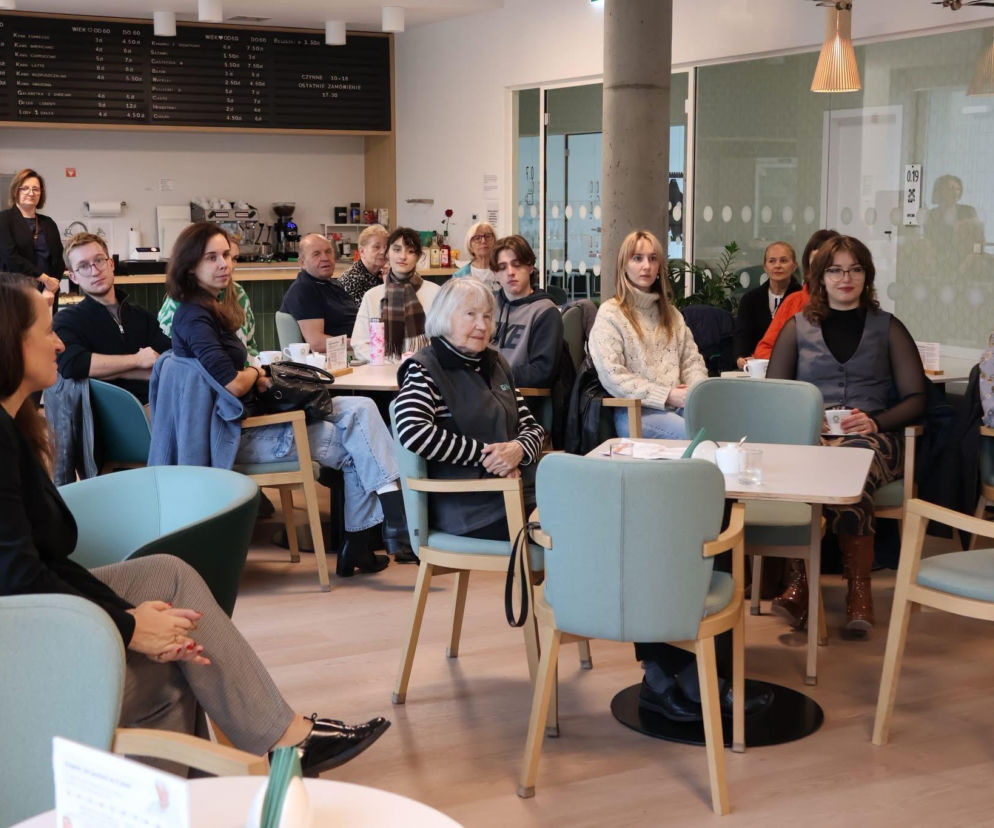 Grupa seniorów i studentów siedzi przy stolikach w jasnej kawiarni CAM. O międzypokoleniowych warsztatach pisze Architektura.