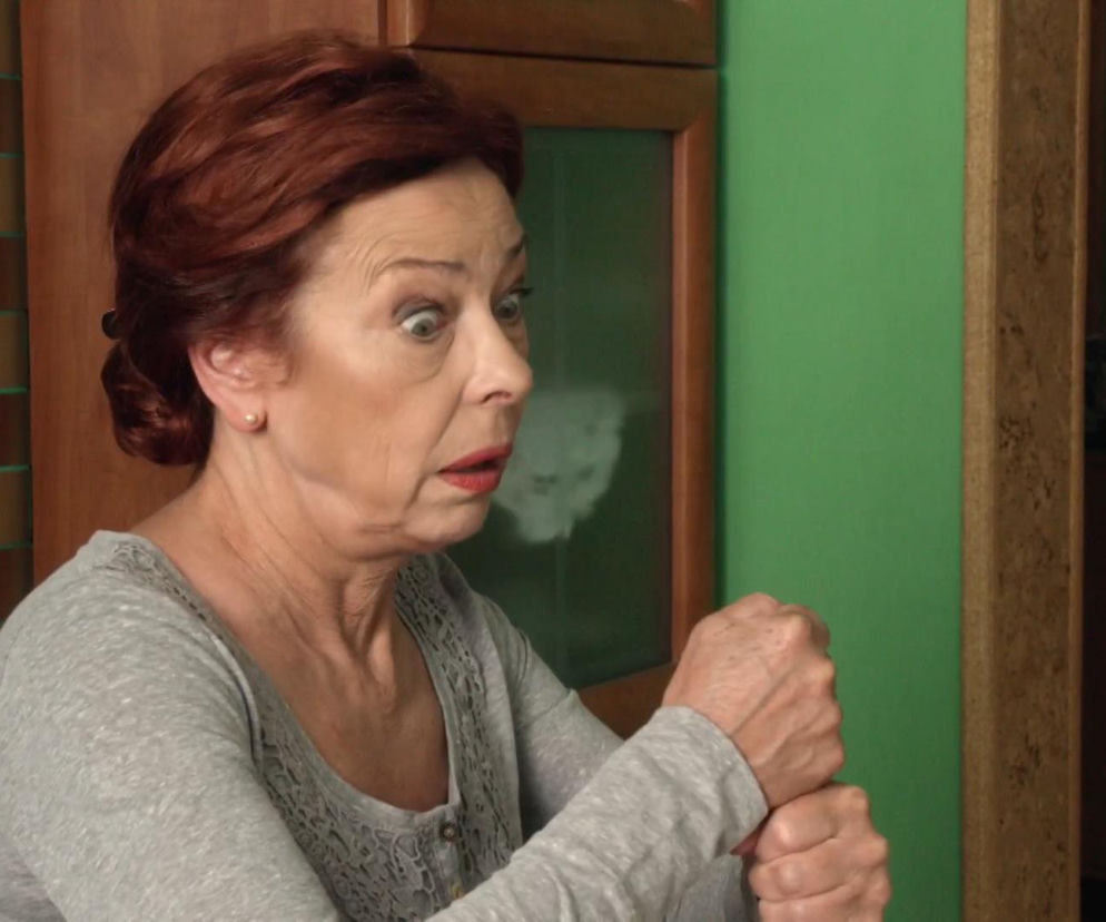 Halina Kozioł z serialu Ranczo, w szarej bluzce z koronką, z zaskoczoną miną i szeroko otwartymi oczami, zaciśniętymi pięściami. Tło stanowi zielona ściana i meble. Więcej o losach bohaterów można przeczytać na naszym portalu.