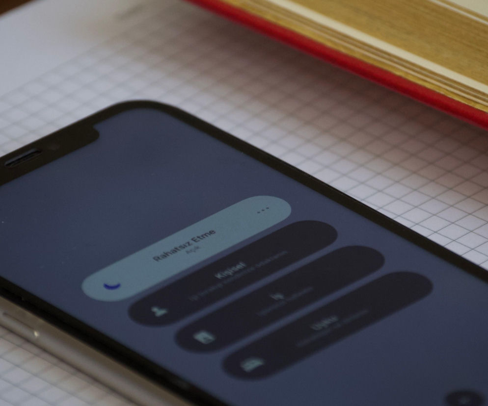 Smartfon leżący na kartce w kratkę obok książki. O zakazie telefonów w szkołach przeczytasz na Eska.