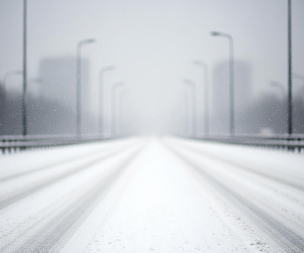 Pusta, zaśnieżona autostrada rozciąga się w centrum kadru, pokryta białym, świeżym śniegiem, z wyraźnie widocznymi, równoległymi śladami kół samochodowych. Po obu stronach drogi biegną czarne barierki ochronne, częściowo zasypane śniegiem. W tle, w gęstej, szarej mgle, widoczne są rozmyte sylwetki latarni ulicznych i niewyraźne zarysy wysokich budynków. Cała scena jest utrzymana w monochromatycznej palecie bieli i szarości, z drobnymi, białymi punktami imitującymi opadający śnieg lub drobinki lodu.