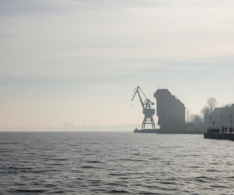 Na pierwszym planie rozciąga się ciemnoszara, falująca tafla wody z drobnymi zmarszczkami odbijającymi jasne światło. Po prawej stronie widoczny jest betonowy brzeg z promenadą, wzdłuż której rozmieszczone są czarne latarnie o klasycznym kształcie, oświetlające ścieżkę, po której spacerują grupy ludzi, stanowiące ciemne sylwetki. Dalej na brzegu, w centrum kadru, stoi duży, zabytkowy żuraw portowy, a za nim monumentalny, ceglany budynek z dwuspadowym dachem i wieżyczką, zanurzony w jasnej, mglistej poświacie. Tło wypełnia zamglony krajobraz miejski z niewyraźnymi zarysami budynków, przechodzący w jasne, niemal jednolite, lekko zachmurzone niebo, przez które widać cienki ślad smugi kondensacyjnej.