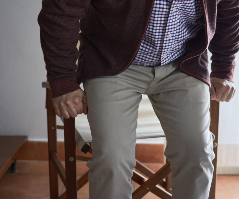 Osoba starsza, prawdopodobnie mężczyzna, z trudem wstająca z drewnianego krzesła, opierająca się dłońmi o jego oparcie. Scena ilustruje codzienne wyzwania seniorów związane z mobilnością, o których więcej przeczytasz na Poradnik Zdrowie.