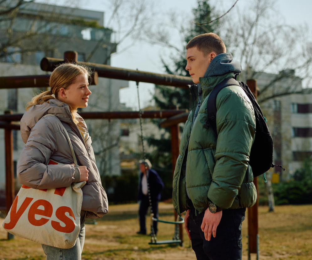 Młoda blondynka w jasnej kurtce i młody mężczyzna w zielonej kurtce rozmawiają na placu zabaw, w tle widać bloki mieszkalne. Scena nawiązuje do serialu Na Wspólnej, o którym więcej można przeczytać na Super Seriale.