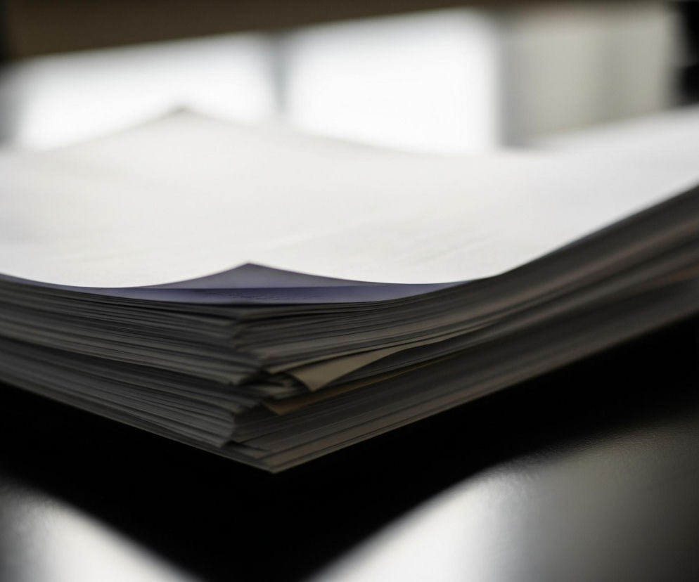 Stos białych kartek papieru leży na czarnym, lśniącym blacie, dominując centrum kadru. Górne krawędzie kartek są lekko zagięte, tworząc falisty kształt i odsłaniając dolne kartki. W tle rozmyte są jasne, białe kształty, prawdopodobnie okna, oświetlające scenę z tyłu, co tworzy silny kontrast z ciemnym blatem. Ciemne cienie papieru oraz odbicia światła są widoczne na błyszczącej powierzchni blatu.