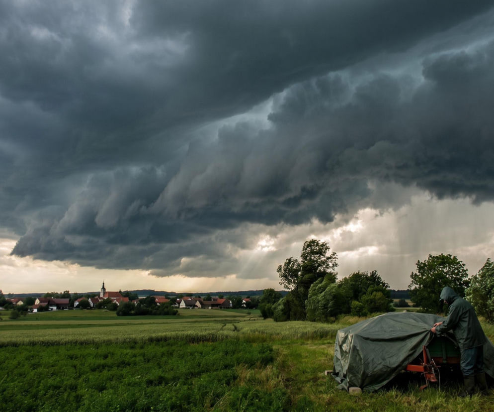 Mężczyzna w płaszczu przeciwdeszczowym zakrywa maszynę plandeką na tle ciemnych chmur burzowych nad wsią, sygnalizując nadejście burzy. Więcej o ostrzeżeniach IMGW przeczytasz na naszym portalu.