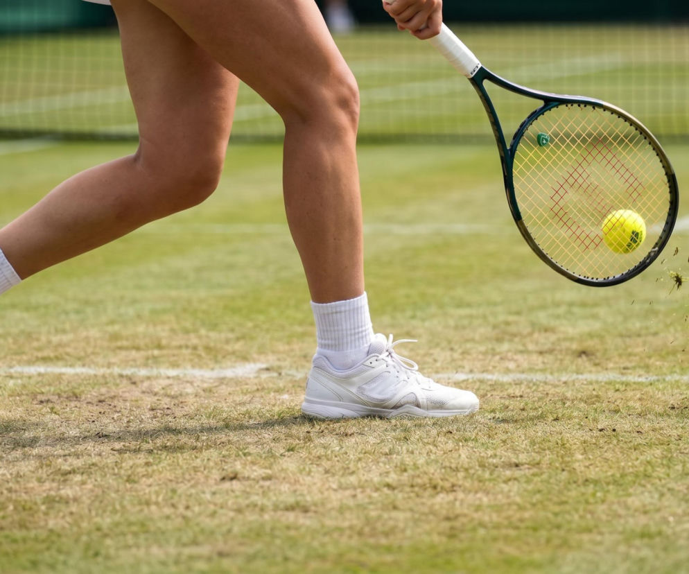 Nogi tenisistki Mai Chwalińskiej w białych butach i skarpetkach na trawiastym korcie, odbijającej żółtą piłkę rakietą, co symbolizuje jej przygotowania do Wimbledonu. Więcej o jej występie na naszym portalu.