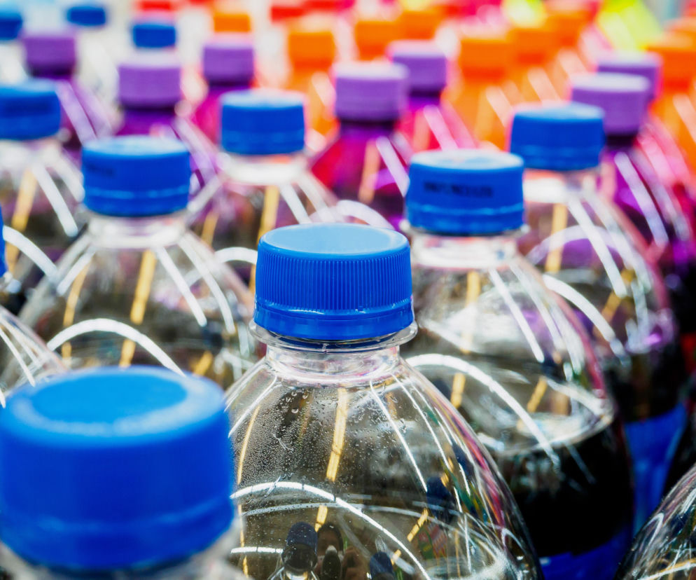Rzędy plastikowych butelek z kolorowymi napojami i niebieskimi zakrętkami. O ich wpływie na dietę przeczytasz na PZ Fit.
