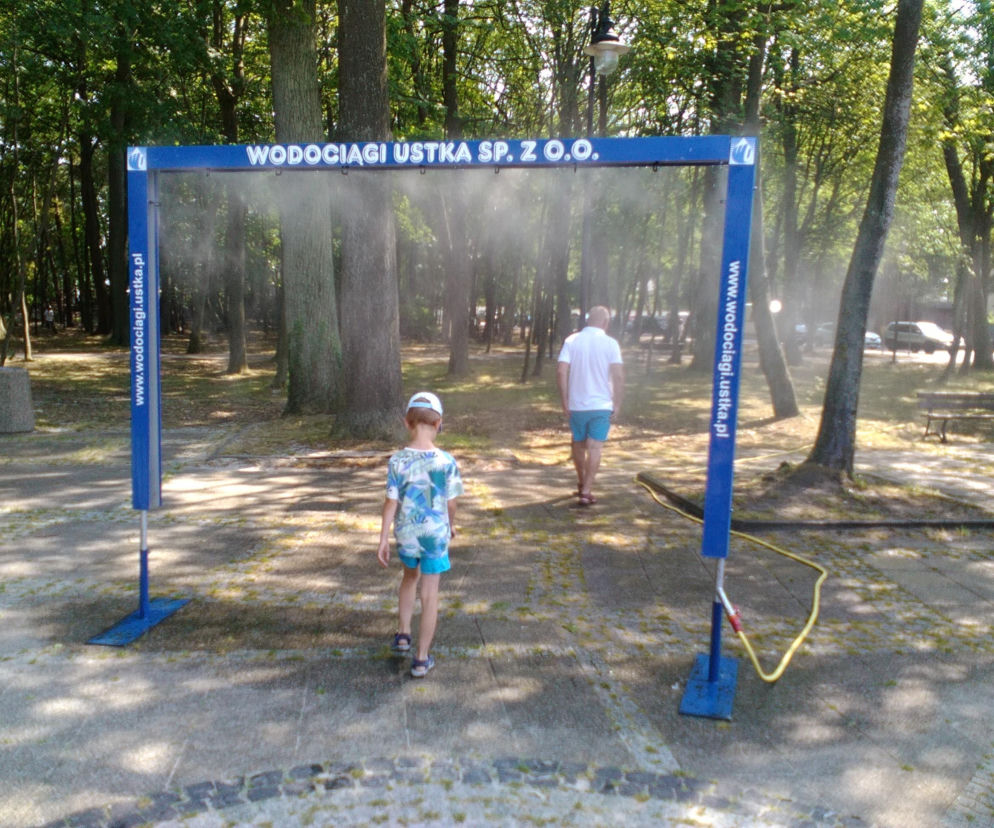 Błękitna kurtyna wodna WODOCIĄGI USTKA SP. Z O.O. zamontowana w parku, z której wydobywa się mgiełka wodna, dająca ochłodę w upalny dzień. Widać idącego mężczyznę i dziecko, którzy przechodzą przez system zraszający. Na naszym portalu znajdziesz informacje o cenach takich kurtyn.