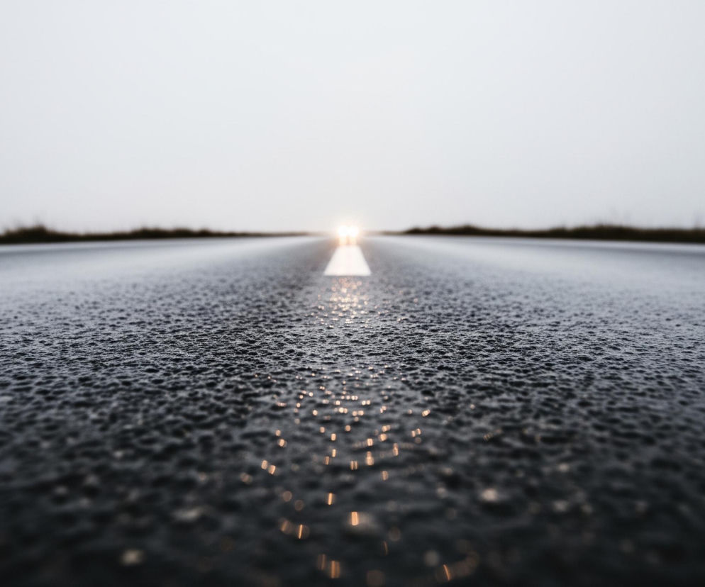 Asfaltowa droga, pokryta licznymi kroplami wody odbijającymi światło, rozciąga się w głąb kadru, tworząc wyraźną perspektywę. Pośrodku niej biegnie biała, ciągła linia, która w oddali łączy się z dwoma jaskrawymi punktami światła, sugerującymi reflektory nadjeżdżającego pojazdu. Po obu stronach drogi widać rozmazane, ciemne pasma roślinności, a nad nimi dominuje jasne, jednolite niebo, co wskazuje na pochmurną pogodę.
