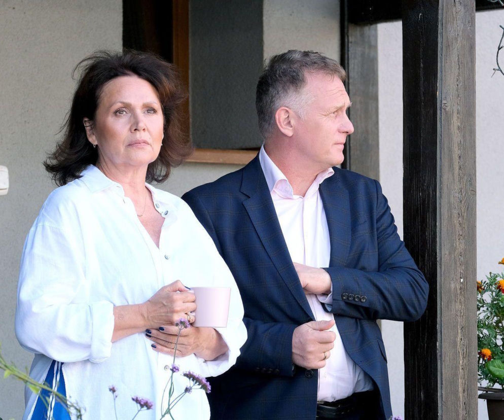 Marysia Rogowska (Małgorzata Pieńkowska) z zatroskaną miną, w białej koszuli i z kubkiem w dłoni, stoi obok Artura Rogowskiego (Robert Moskwa) w granatowej marynarce i jasnej koszuli. Obydwoje patrzą w prawo, a obok nich widoczne są rośliny i elementy drewnianej konstrukcji, co sugeruje scenę z serialu M jak miłość. Więcej informacji o serialu znajdziesz na Super Seriale.