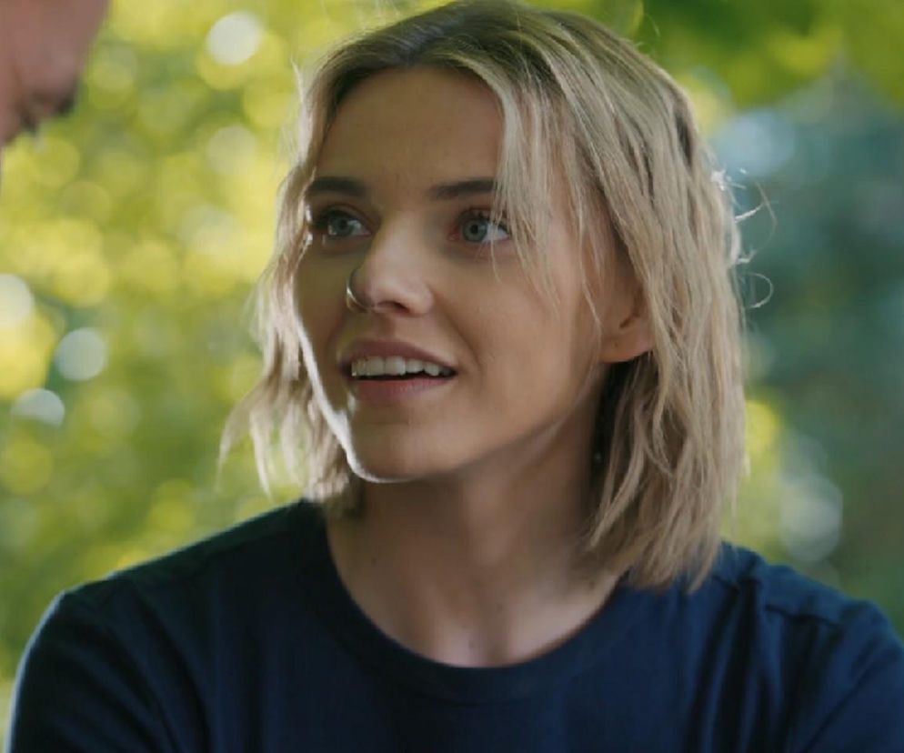 Zmysłowa Oliwia Drożdżyk jako Janka z blond włosami, niebieskimi oczami i kolczykiem w nosie, patrząca z uśmiechem na Rafała Cieszyńskiego grającego Mariusza. Para z serialu „Barwy szczęścia w romantycznej scenie, o której przeczytasz więcej na portalu Super Seriale.