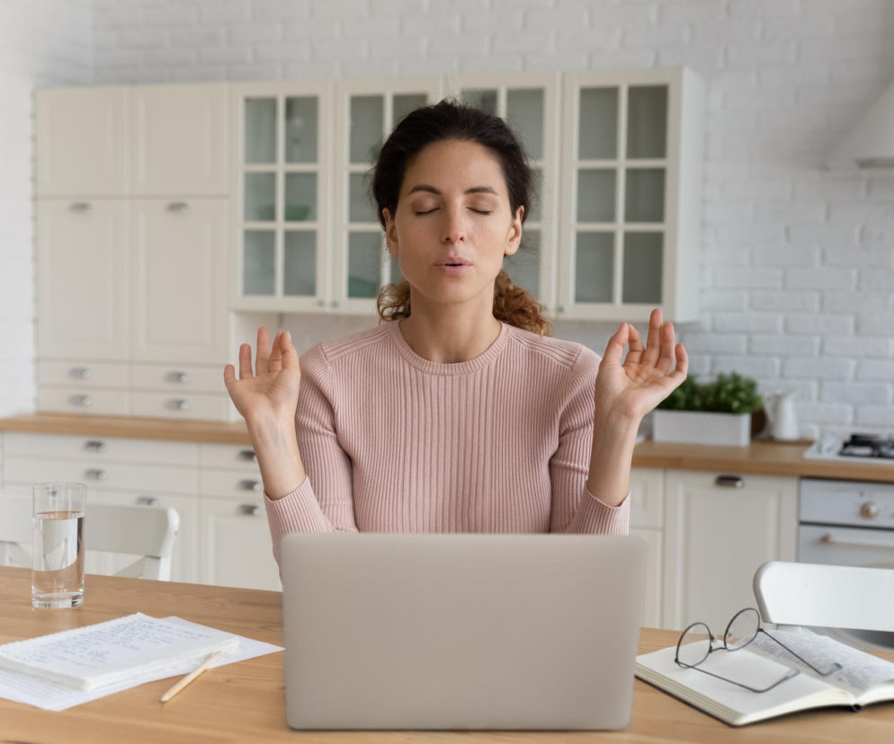 Kobieta z zamkniętymi oczami i dłońmi ułożonymi w geście medytacji, siedząca przy stole z laptopem. Opiera się na technice oddechowej, która redukuje stres. Więcej o metodzie szybszej niż medytacja na Poradnik Zdrowie.