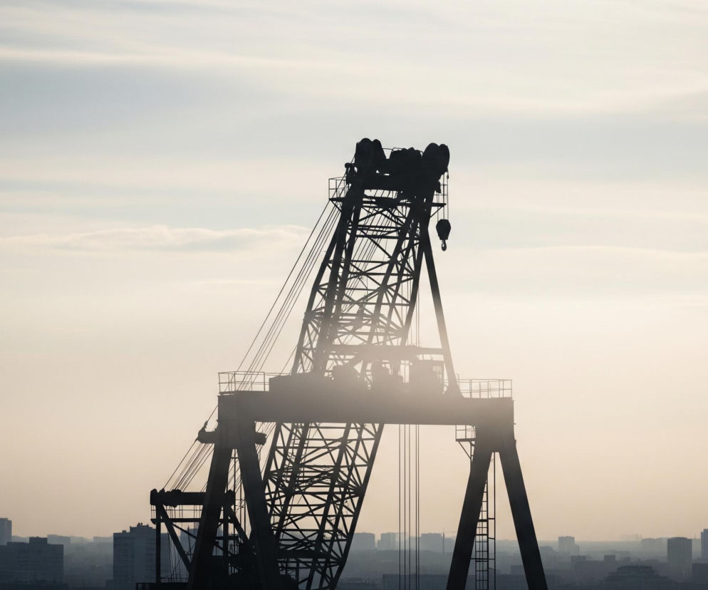 Sylwetka potężnego dźwigu portowego, wykonana w ciemnym kolorze, dominuje nad horyzontem, zajmując centralną część kadru. Za dźwigiem rozciąga się jasne, zamglone niebo o odcieniach błękitu i jasnego beżu, z widocznymi poziomymi smugami chmur. U dołu obrazu, pod dźwigiem, widoczna jest panoramiczna, zamglona sylwetka miasta złożona z licznych wieżowców i budynków, których kształty rozmywają się w oddali, tworząc wrażenie głębi i perspektywy. Całość kompozycji oświetla miękkie, rozproszone światło, tworząc atmosferę spokoju i monumentalności.