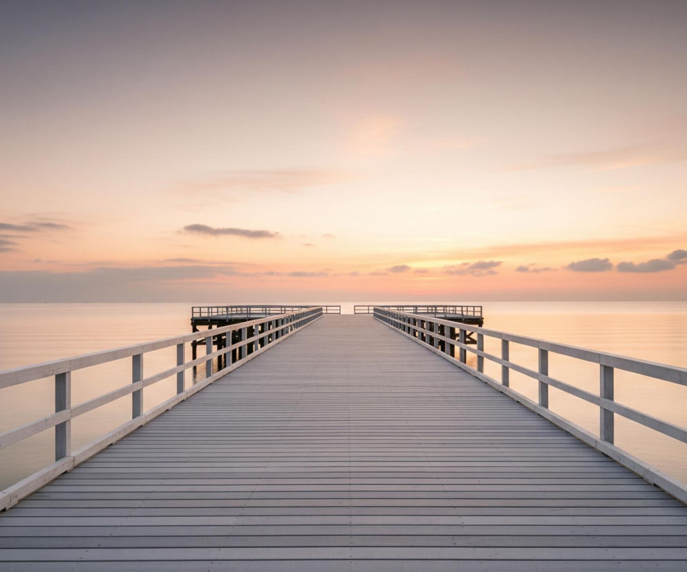Długie, drewniane molo, pomalowane na jasny odcień szarości, rozciąga się w głąb spokojnej wody, dominując centralną część obrazu. Po obu stronach molo biegną białe, drewniane balustrady, składające się z dwóch poziomych belek i pionowych słupków. Horyzont jest nisko położony, a niebo nad nim prezentuje gradient kolorów od jasnego błękitu na górze, poprzez delikatne róże i pomarańcze w centrum, do jaśniejszych odcieni w pobliżu horyzontu. Kilka małych, szarych chmur unosi się na niebie, a na wodzie widoczne są subtelne refleksy światła.