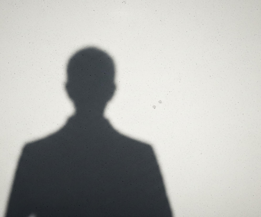 Ciemny cień ludzkiej sylwetki na jasnej, ziarnistej powierzchni. Cień przedstawia osobę w pozycji frontalnej, z wyraźnie zarysowanymi ramionami, tułowiem i głową, przypominającą kształt męskiej postaci w marynarce. Tło to jednolita, jasnoszara płaszczyzna, pokryta drobnymi, ciemniejszymi plamkami, rozrzuconymi nierównomiernie po całej powierzchni. Cień znajduje się po lewej stronie kadru, pozostawiając dużą pustą przestrzeń po prawej.