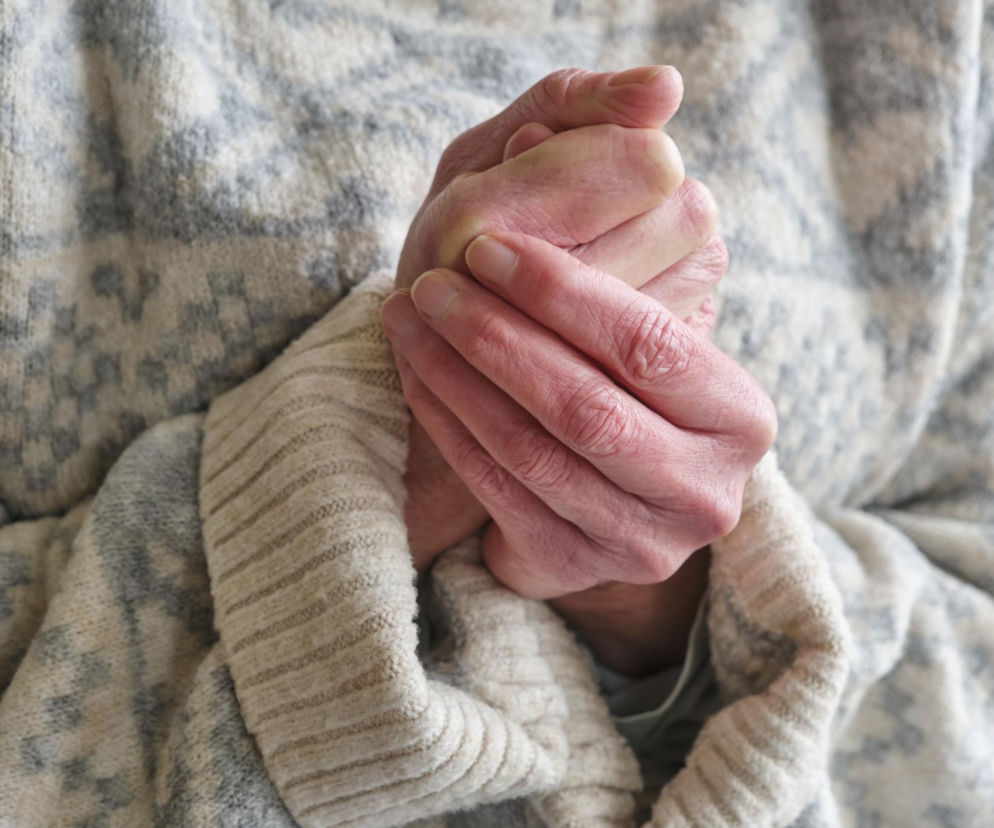 Osoba ogrzewająca zmarznięte dłonie w rękawach kremowo-szarego swetra, co symbolizuje problem zimnych kończyn. Na Poradnik Zdrowie można przeczytać o tym, kiedy zimne dłonie i stopy są objawem choroby, wymagającym konsultacji lekarskiej.