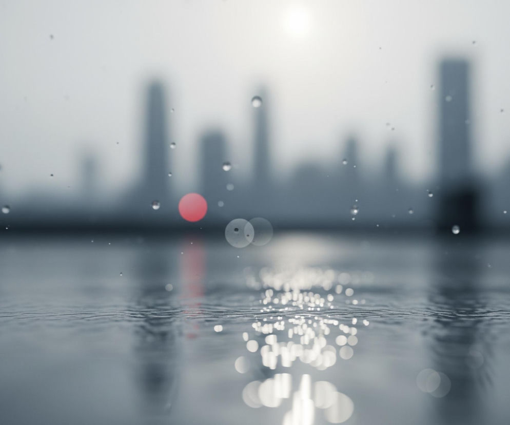 Dolna połowa obrazu ukazuje szarą powierzchnię wody z delikatnymi falami i licznymi, jasnymi, okrągłymi refleksami światła odbijającymi się od niej, szczególnie w prawej części. Powyżej, w górnej połowie, widać rozmyte tło przedstawiające sylwetki wysokich budynków w odcieniach szarości, z pojedynczym, jaskrawoczerwonym punktem światła pośrodku. Na pierwszym planie, w powietrzu, widoczne są pojedyncze krople wody, rozrzucone nieregularnie na tle nieba, oraz większe, wyraźne krople na dole obrazu. W górnej części obrazu, pośrodku, znajduje się jasna, rozmyta plama, prawdopodobnie słońce, a cała scena utrzymana jest w chłodnej palecie barw, głównie szarości i błękitu.