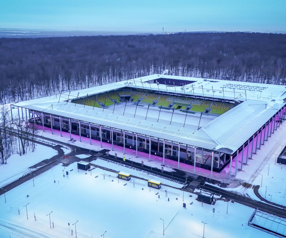 Nowy Stadion Miejski Arena Katowice nocą w zimowej scenerii, z zielono-żółtymi krzesełkami i podświetloną kolumnadą w odcieniach fioletu. Obiekt prezentuje minimalistyczną architekturę i jest nominowany do Stadium of the Year 2025, o czym możesz przeczytać na Architektura Murator Plus.