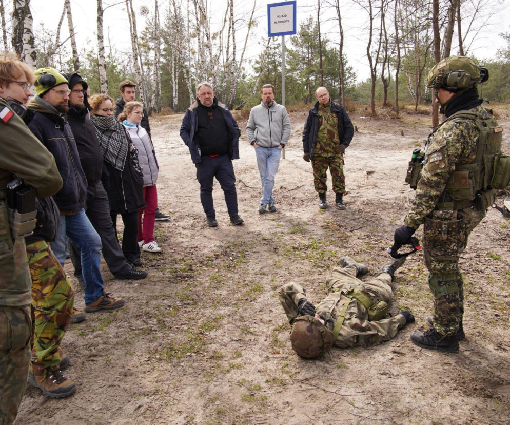 Grupa cywilów obserwuje żołnierza udzielającego pomocy leżącej osobie na poligonie. Więcej na SE Portal Obronny.