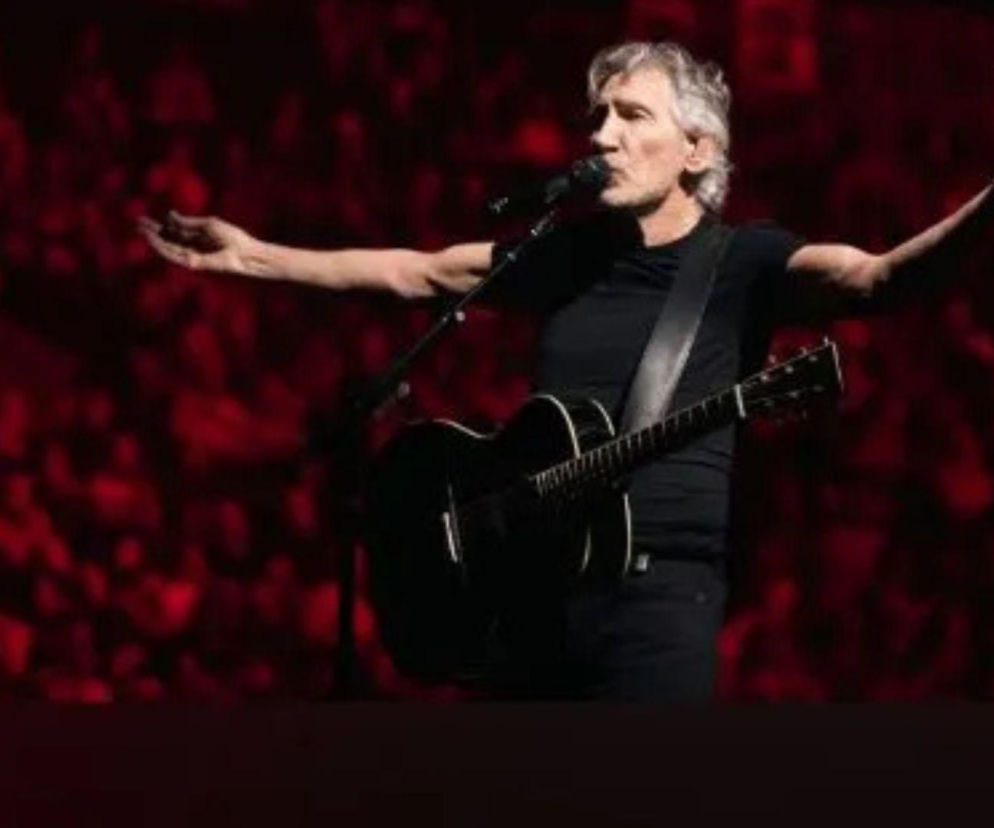 Roger Waters na scenie, ubrany na czarno, z akustyczną gitarą, śpiewa do mikrofonu z rozłożonymi rękami na tle czerwonego światła. Więcej o nowej wersji klasyków Pink Floyd przeczytasz na naszym portalu.